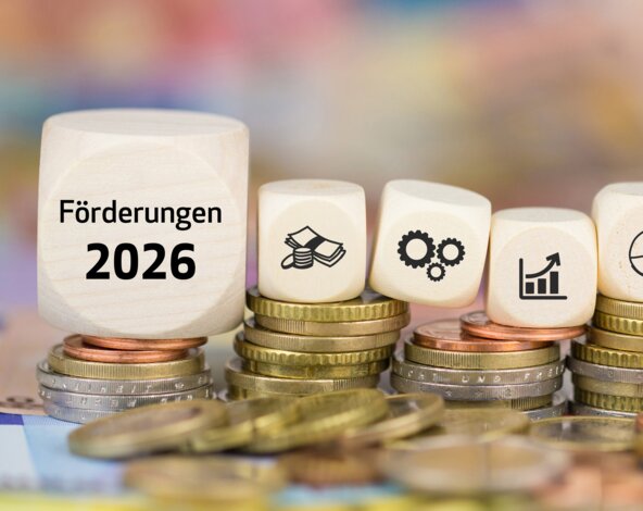 Weiße Würfel mit der Aufschrift 'Förderungen 2026' und Finanz-Icons auf gestapelten Euro-Münzen – Ratgeber zu staatlichen Zuschüssen für klimafreundliche Heizungen