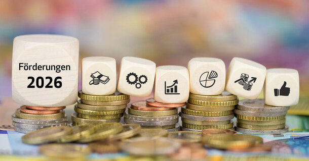 Weiße Würfel mit der Aufschrift 'Förderungen 2026' und Finanz-Icons auf gestapelten Euro-Münzen – Ratgeber zu staatlichen Zuschüssen für klimafreundliche Heizungen
