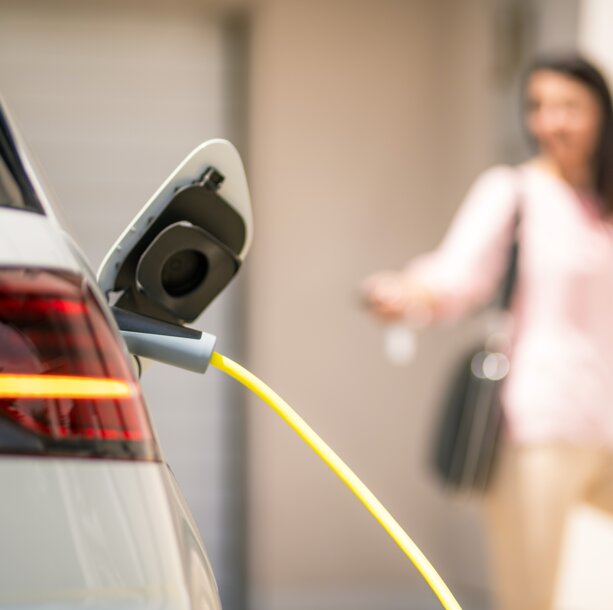 Ein Elektroauto wird über ein gelbes Kabel geladen, im Hintergrund eine Frau – Clevere Nutzung der PV-Anlage, um das E-Auto nach Feierabend mit Solarstrom zu laden