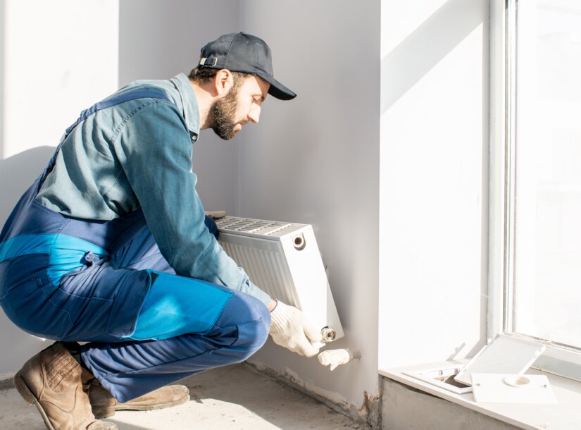 Ein Handwerker in blauer Arbeitskleidung montiert einen Heizkörper unter einem Fenster – Ratgeber zum neuen Heizungsgesetz (GEG) 