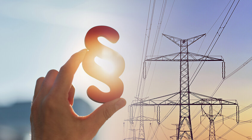 Eine Hand hält ein Paragraph-Zeichen vor Hochspannungsmasten im Sonnenlicht – Informationen zu Gesetzen und staatlichen Entlastungen bei den Energiepreisen