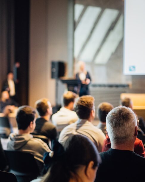 Blick aus dem Publikum auf eine Rednerin bei einer Fachkonferenz – Expertenvorträge und Diskussionsrunden für Immobilienverwalter