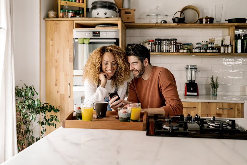 Lächelndes Paar in der Küche schaut auf ein Smartphone – Tarifrechner für den optimalen Erdgastarif starten