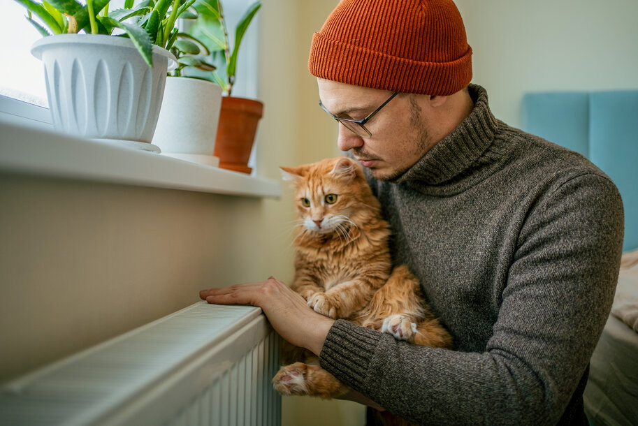 Mann mit Katze auf dem Arm fühlt die Temperatur am Heizkörper – Tipps zum effizienten Heizen und Heizkosten sparen
