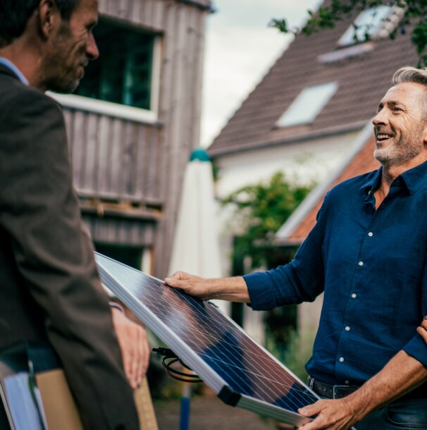 Ein lächelnder Kunde und ein Berater halten gemeinsam ein Solarmodul vor einem Haus – MONTANAs Fokus auf eine nachhaltige Zukunft und intelligente Energielösungen