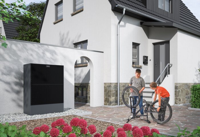 Eine moderne Wärmepumpe an der Außenwand eines Wohnhauses, im Hintergrund reparieren zwei Kinder ein Fahrrad – MONTANA Wärmepumpen-Lösungen für Bestandsgebäude