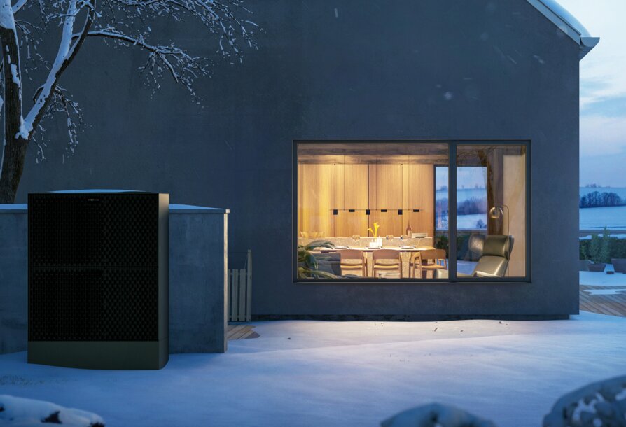 Außeneinheit einer Wärmepumpe vor einem beleuchteten Wohnhaus im Winter – Nutzung von günstigem Wärmestrom