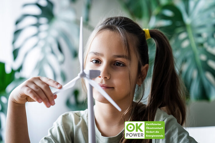 Mädchen betrachtet interessiert ein Modell-Windrad; ok-power Ökostrom Zertifikat im Vordergrund