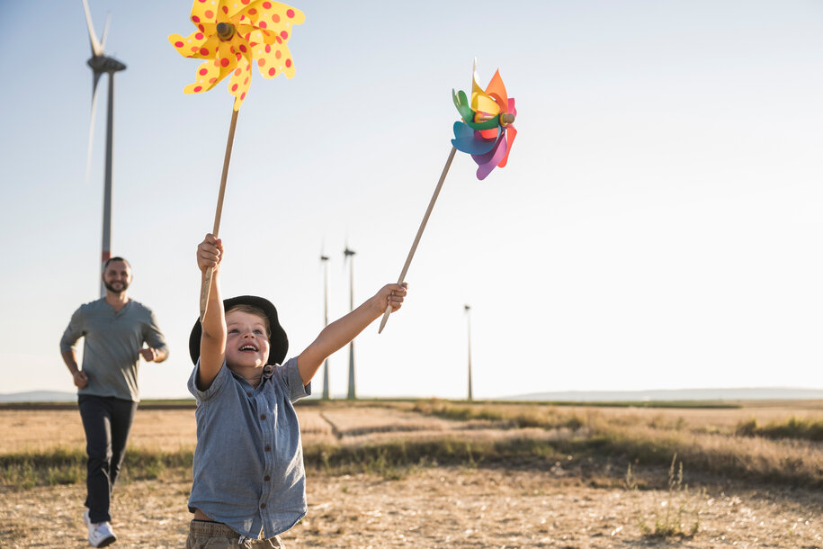 Kind spielt mit bunten Windrädern auf einem Feld, im Hintergrund Windkraftanlagen