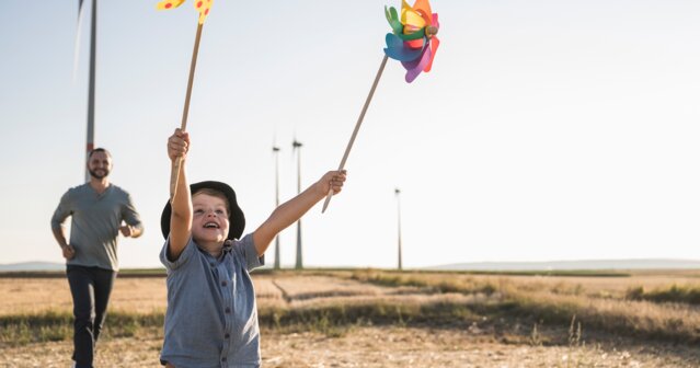 Kind spielt mit bunten Windrädern auf einem Feld, im Hintergrund Windkraftanlagen