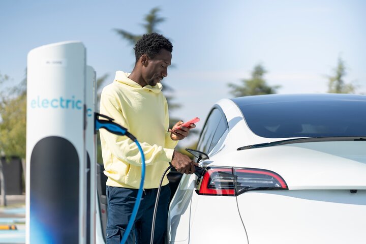 Fahrer lädt sein E-Auto an einer Stromladesäule mit Ladekabel und Smartphone in der Hand.