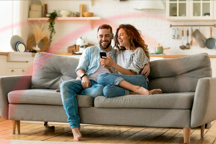 Lachendes Paar auf dem Sofa schaut auf ein Smartphone – attraktive MONTANA Stromtarife