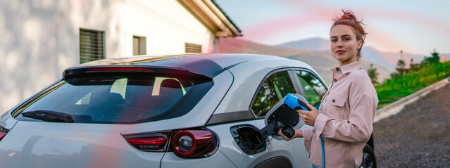 Frau mit Ladekabel am Elektroauto vor Bergkulisse – der günstige MONTANA Autostrom-Tarif für Ihr E-Auto