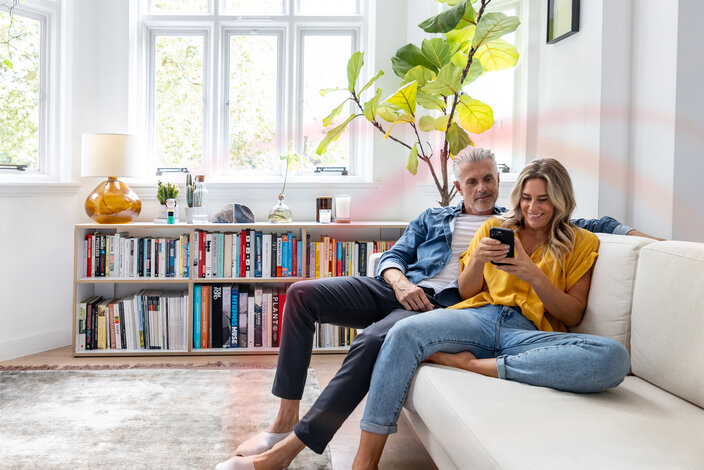 Paar im hellen Wohnzimmer wählt Gastarif auf dem Smartphone – Anmeldung für Neukunden bei Neueinzug