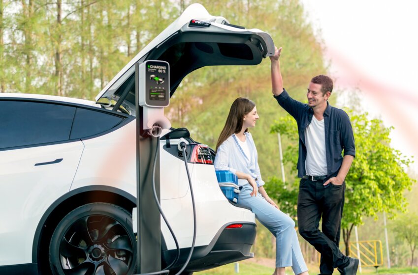 Ein Paar lächelt beim Laden ihres E-Autos an einer Ladestation im Grünen – Unterwegs in Europa mit der MONTANA Ladekarte für Elektromobilität