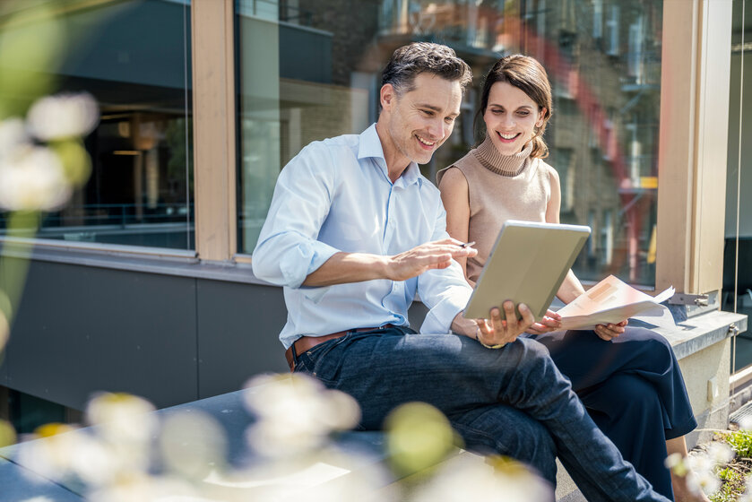 Geschäftspartner sitzen im freien und schauen lächelnd in ein Tablet – Newsletter für die Wohnungswirtschaft