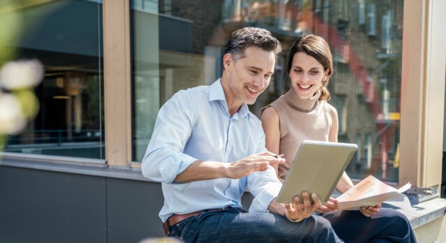 Geschäftspartner sitzen im freien und schauen lächelnd in ein Tablet – Newsletter für die Wohnungswirtschaft