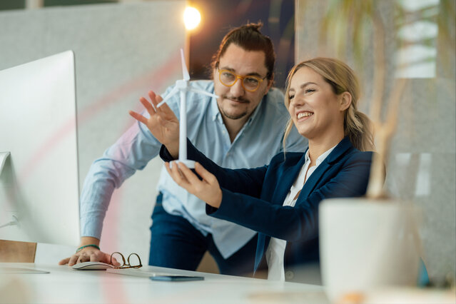 Zwei Geschäftspartner betrachten lächelnd ein Modell eines Windrads – Jetzt individuellen Strompreis im MONTANA Gewerbestrom-Tarifrechner berechnen